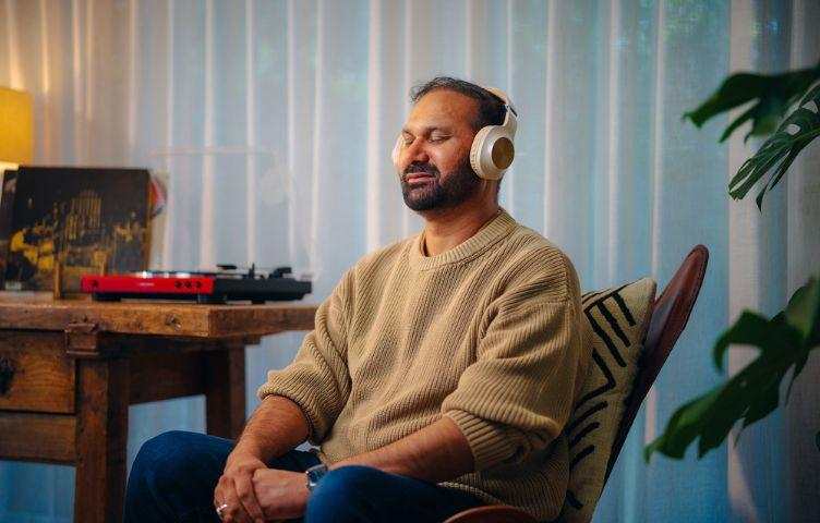 A man sits on an armchair in his living room. He wears a pair of headphones while listening to a podcast.