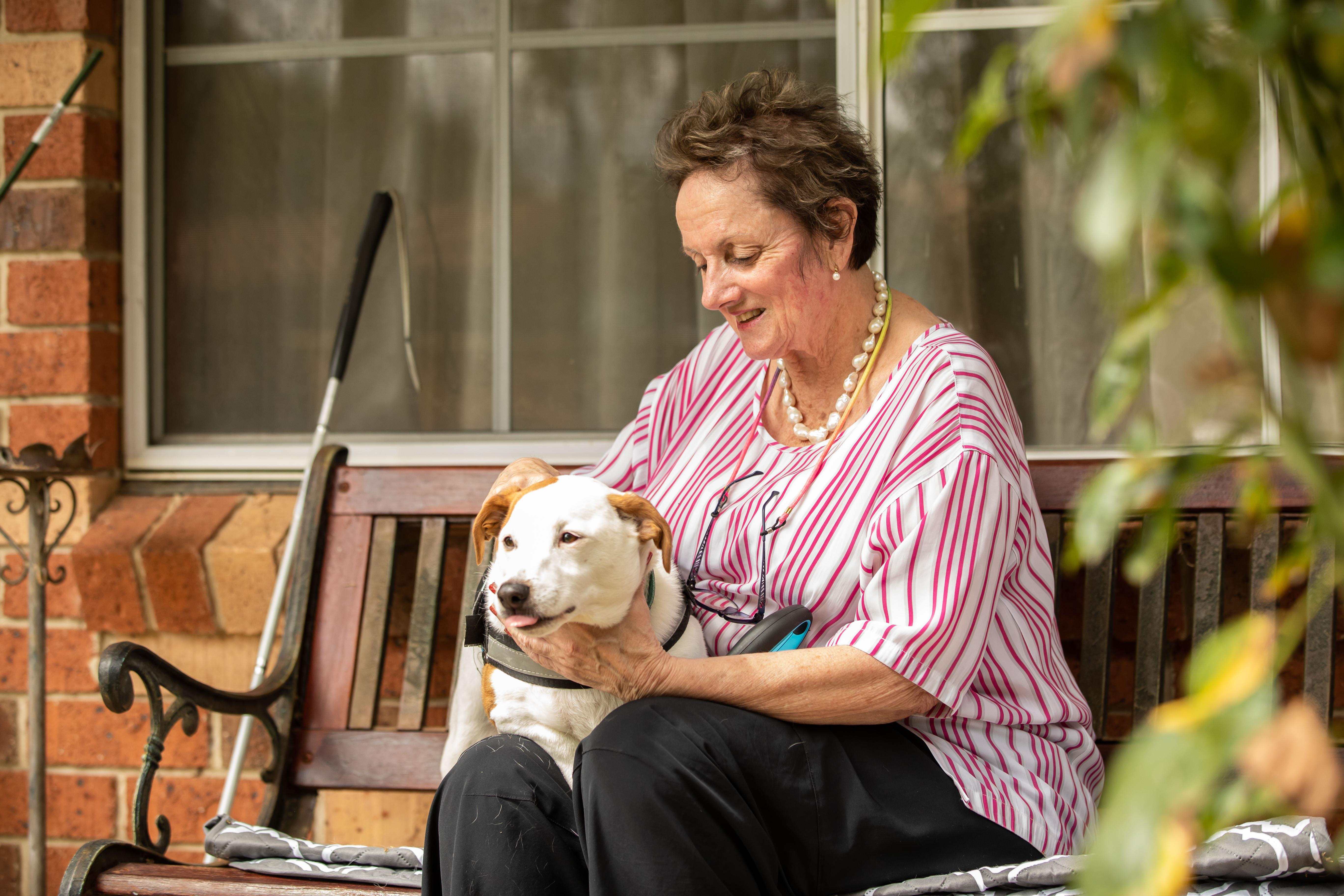 A smiling older woman sits on a bench, petting a white and brown dog. A white cane leans beside her.
