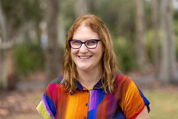 A headshot of Dr Katie Butler. She is staring into the camera, smiling. She is wearing black square framed glasses and a very colourful shirt.
