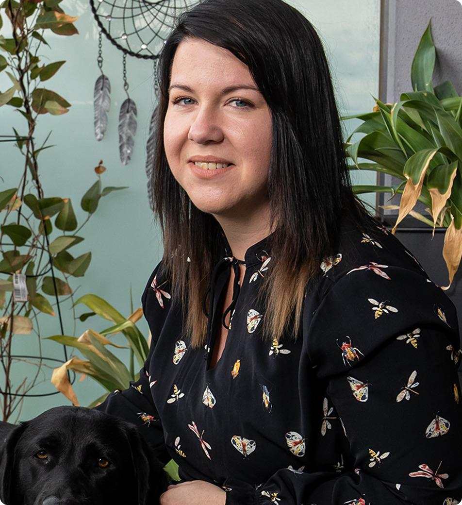 Brittnee, a young woman with long dark hair, sits outdoors with a black guide dog, surrounded by plants and a dreamcatcher.