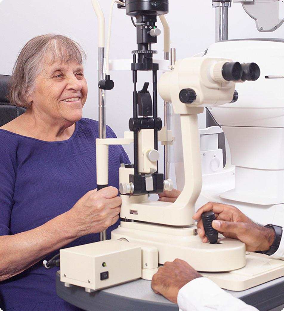 An older woman smiles while having an eye exam with a slit lamp. A healthcare professional adjusts the machine.