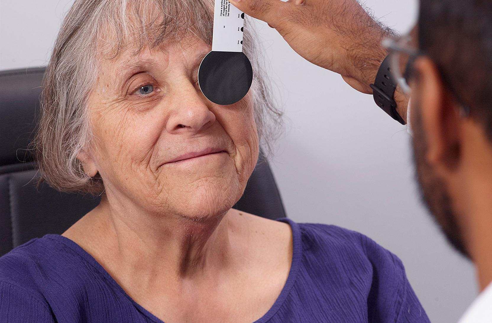 An older woman with grey hair and a warm smile undergoes an eye examination. A healthcare professional holds an occluder over one of her eyes.