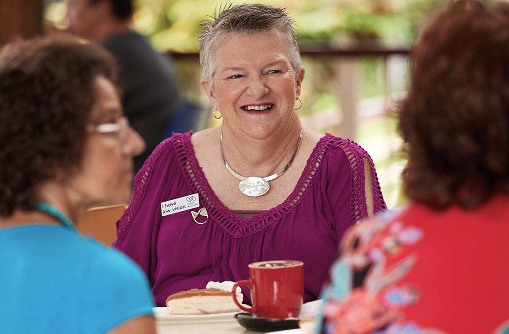 A woman in a purple top with a "low vision" badge smiles while chatting at a café table with coffee and cake.