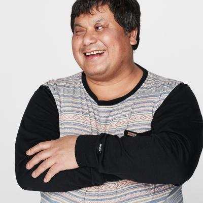 A young man with short dark hair smiles and crosses his arms. He wears a striped shirt with black sleeves against a plain white background.
