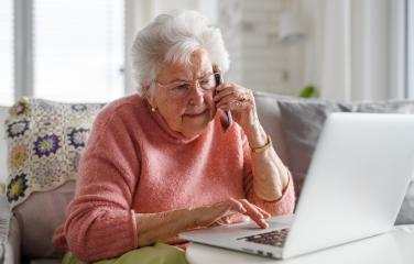 An elderly lady is sitting in front of a laptop. She also looks to be listening to someone speaking on her mobile phone.