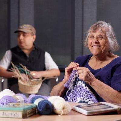 An older woman in a purple top knits with purple and white yarn. A man in the background weaves a basket.