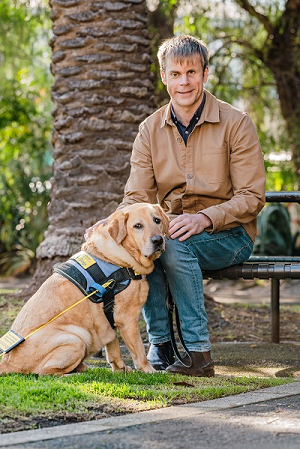 Sam Byrne is sitting on a park bench under a tree, with a yellow Seeing Eye Dog sitting on the ground next to him.
